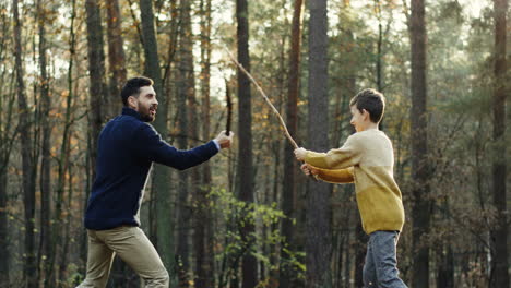 Cheerful-Caucasian-father-and-little-son-playing-in-the-forest-with-sticks-like-they-fighting-with-swords