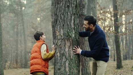 Joyful-caucasian-father-and-son-playing-in-the-wood,-hiding-behind-a-tree-trunk-and-trying-to-scape