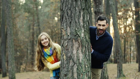Portrait-shot-of-Caucasian-handsome-dad-and-his-cute-little-girl-looking-at-camera-behind-a-tree-trunk-in-the-forest