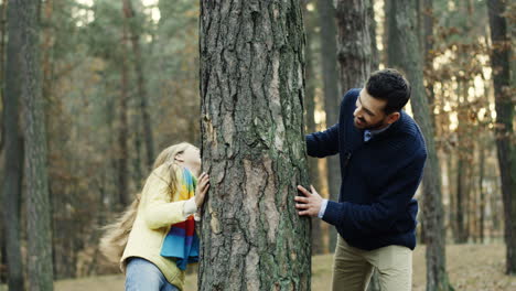 Joyful-caucasian-father-and-daughter-playing-in-the-wood,-hiding-behind-a-tree-trunk-and-looking-at-each-other-from-different-sides