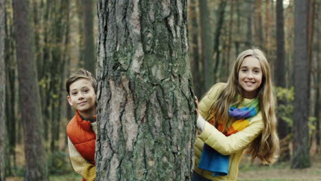 Portrait-shot-of-cheerful-cute-boy-and-girl-looking-at-camera-behind-a-tree-trunk-in-the-forest