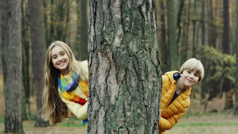 Portraitaufnahme-Von-Fröhlichen,-Süßen-Jungen-Und-Mädchen,-Die-Hinter-Einem-Baumstamm-Im-Wald-In-Die-Kamera-Schauen