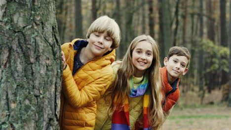 Portrait-of-two-cute-boys-and-one-girl-looking-at-camera-behind-a-tree-trunk-in-the-forest