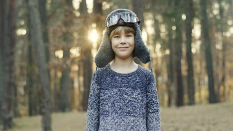 Kleiner-Kaukasischer-Junge,-Der-Davon-Träumt,-Ein-Pilot-Zu-Sein,-Der-Spaß-Hat-Und-Mit-Hut-Und-Spezialbrille-Läuft-Und-Im-Wald-In-Die-Kamera-Lächelt