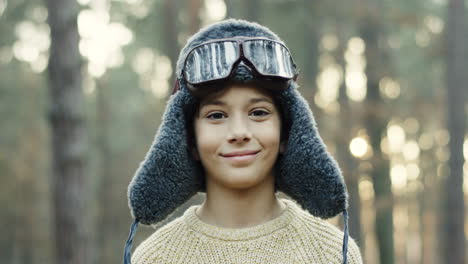 Close-up-view-of-little-Caucasian-boy-dreamer-of-being-a-pilot-in-hat-and-special-glasses-smiling-at-the-camera-in-the-forest
