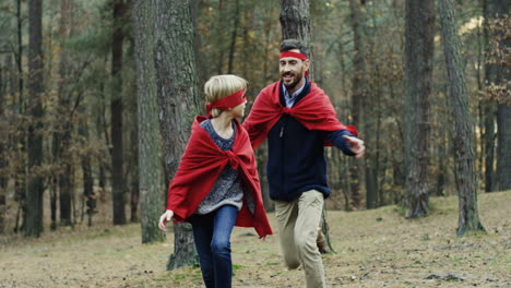 Happy-Caucasian-dad-trying-to-cath-his-small-son-while-they-playing-being-superheroes-with-red-capes-in-the-forest