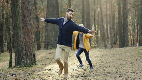 Caucasian-happy-father-and-his-little-son-running-in-the-forest-with-hands-open-imitating-an-airplane-flying