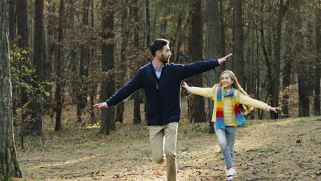 Caucasian-happy-father-and-his-daughter-running-in-the-forest-with-hands-open-imitating-an-airplane-flying