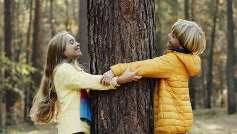 Joyful-caucasian-sister-and-brother-playing-in-the-wood,-hiding-behind-a-tree-trunk-and-looking-at-each-other-from-different-sides