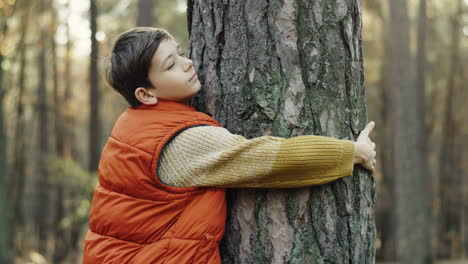 Side-view-of-a-cute-little-teen-boy-hugging-a-tree-trunk-with-eyes-closed-in-the-forest