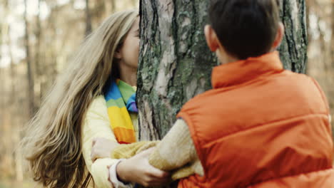 Close-up-view-of-caucasian-sister-and-brother-playing-in-the-wood,-hiding-behind-a-tree-trunk-and-looking-at-each-other-from-different-sides