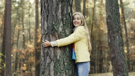 Side-view-of-a-cute-teen-girl-hugging-a-tree-trunk-with-eyes-closed-in-the-forest