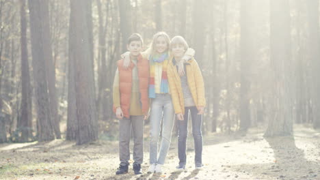 Distant-view-of-joyful-kids,-girl-and-two-boys,-hugging-and-smiling-at-camera-in-the-forest-on-a-sunny-day