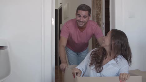 Funny-woman-sitting-inside-carton-box-while-her-boyfriend-pushing-her-through-the-corridor-of-the-new-house