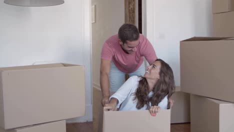 Funny-woman-sitting-inside-carton-box-while-her-boyfriend-pushing-her-through-the-corridor-of-the-new-house