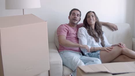 Happy-Caucasian-couple-sitting-on-sofa,-talking-and-hugging-on-moving-day-in-new-house