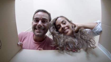 Young-happy-couple-opening-cardboard-box-after-relocation,-looking-inside-and-smiling