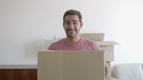 Portrait-of-happy-young-man-standing-in-the-living-room-of-his-new-house,-holding-a-cardboard-box-and-smiling-at-the-camera