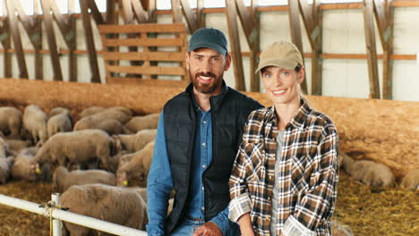 Portrait-of-happy-young-Caucasian-married-couple-of-farmers-looking-at-each-other-and-smiling-at-camera-while-sitting-in-stable-with-sheep-flock