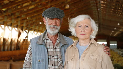 Portrait-of-happy-old-Caucasian-married-couple-of-farmers-looking-and-smiling-at-camera-while-standing-in-stable-with-sheep-flock