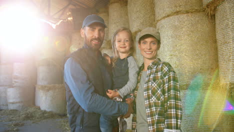 Portrait-of-happy-Caucasian-family-of-farmers-with-little-girl-standing-at-stable-with-hay-stocks-and-smiling-at-camera