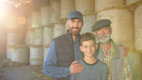 Teen-boy-with-her-father-and-grandfather-standing-in-stable-with-hay-stocks-and-smiling-at-camera