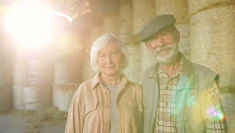 Portrait-of-happy-old-Caucasian-married-couple-of-farmers-looking-and-smiling-at-camera-while-standing-in-stable-with-hay-stocks