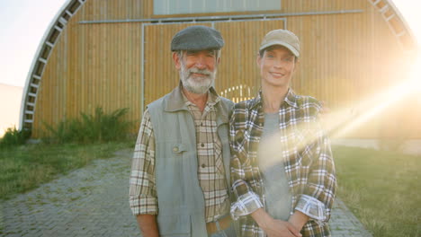 Portrait-of-gray-haired-Caucasian-old-man-with-young-woman-standing-outdoor-at-stable-and-smiling-at-camera-on-a-sunny-day