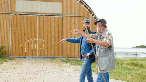 Side-view-of-old-father-and-son-farmers-walking-and-talking-outside-the-stable