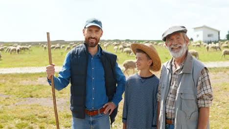 Grandfather,-father-and-grandson-farmers-looking-at-camera-in-green-field-while-the-sheep-flock-is-grazing