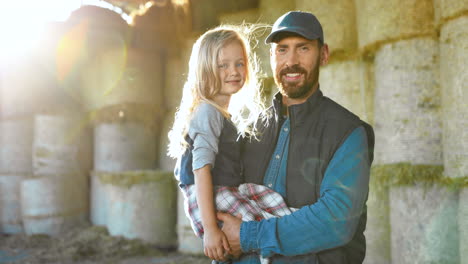 Portrait-of-Caucasian-young-father-holding-on-hands-his-cute-little-daughter-and-smiling-at-camera-in-stable-with-hay-stocks