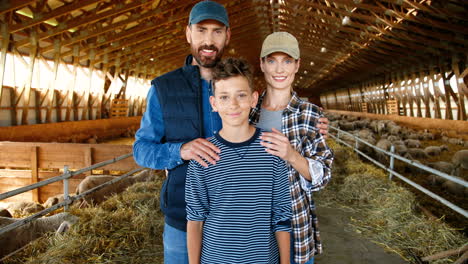 Portrait-of-Caucasian-happy-mother-and-father-with-teenage-son-smiling-at-camera-in-stable-wih-sheep-flock