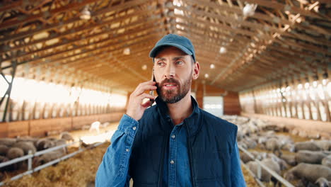 Close-up-view-of-young-Caucasian-man-in-cap-walking-in-stable-with-sheep-flock-and-talking-on-mobile-phone