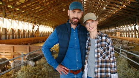Camera-zooming-on-caucasian-young-couple-of-farmers-looking-at-camera-in-a-stable-with-sheep-flock