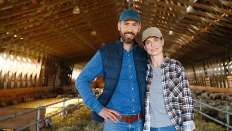 Close-up-view-of-caucasian-young-couple-of-farmers-looking-at-camera-in-a-stable-with-sheep-flock