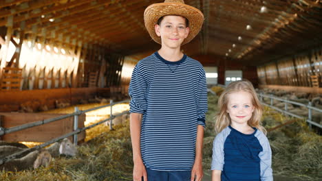 Portrait-of-Caucasian-teen-boy-in-hat-and-his-little-sister-looking-at-camera-in-stable-with-sheep-flock
