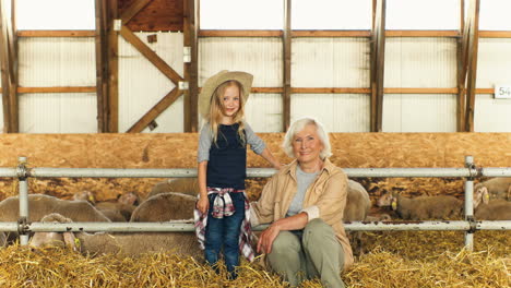 Portrait-of-Caucasian-grandmother-and-her-little-granddaughter-smiling-at-camera-while-sitting-together-in-stable-with-sheep-flock