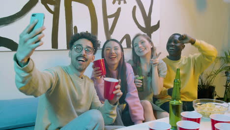 Funny-multiethnic-young-friends-taking-a-selfie-video-while-sitting-on-sofa-and-having-a-beer-party-at-home