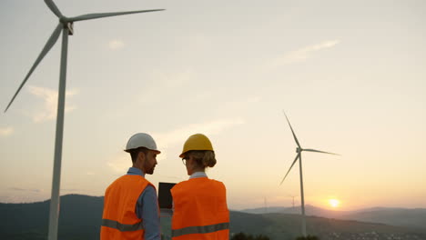 Rear-view-of-a-man-and-woman-in-helmets-discussing-something-at-the-huge-windmills-turbines