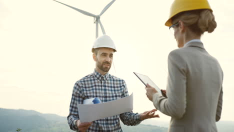 Caucasian-male-engineer-wearing-a-helmet-holding-blueprints-and-talking-with-a-female-colleage-at-the-huge-windmills-turbines