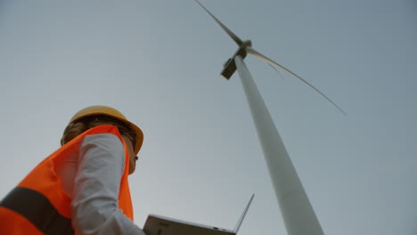 Lower-view-of-a-caucasian-female-engineer-wearing-a-helmet-and-using-laptop-under-the-huge-windmill-turbine-and-watching-it-working-and-producing-energy