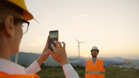 Caucasian-female-engineer-wearing-a-helmet-taking-a-photo-to-her-colleage-with-smartphone-at-the-huge-windmills-turbines-at-sunset