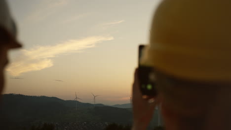 Rückansicht-Einer-Kaukasischen-Arbeiterin,-Die-Einen-Helm-Trägt-Und-Mit-Ihrem-Smartphone-Ein-Foto-Der-Windmühlenturbinen-Macht,-Die-Sich-Bei-Sonnenuntergang-Drehen