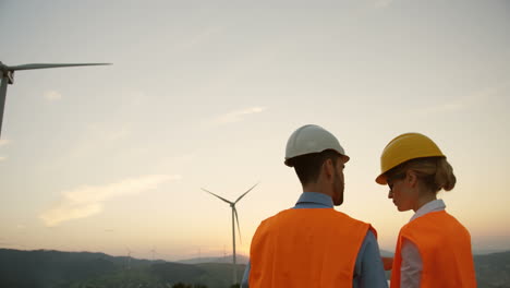 Rear-view-of-two-Caucasian-male-and-female-engineers-in-helmets-talking-about-the-big-windmills-turbines