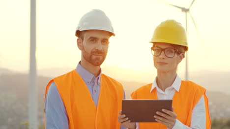 Caucasian-male-and-female-engineers-in-helmets-and-uniforms-using-tablet-and-talking-at-wind-station-of-renewable-energy,-then-they-smile-at-the-camera