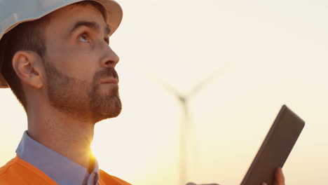 Close-up-view-of-caucasian-male-engineer-in-helmet-and-uniform-using-tablet-at-wind-station-of-renewable-energy
