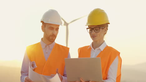 Caucasian-male-and-female-engineers-in-helmet-and-uniform-using-laptop-and-looking-at-blueprints-at-wind-station-of-renewable-energy