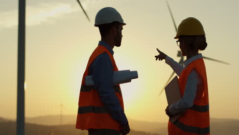 Caucasian-male-and-female-engineers-in-helmet-and-uniform-talking-while-deciding-something-about-operation-of-the-windmills-at-station-of-renewable-energy