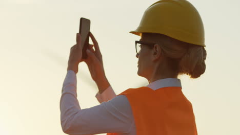 Side-view-of-caucasian-female-worker-wearing-a-helmet-taking-a-photo-with-her-smartphone-of-the-windmills-turbines-spinning-at-sunset