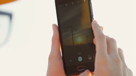 Nahaufnahme-Des-Smartphone-Bildschirms-Mit-Fotos-Der-Riesigen-Windmühlen-Turbinen-Bei-Sonnenuntergang